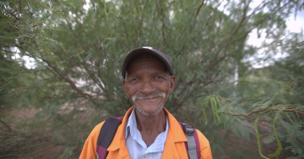 Man wearing an orange jacket and cap standing in front of messquite trees